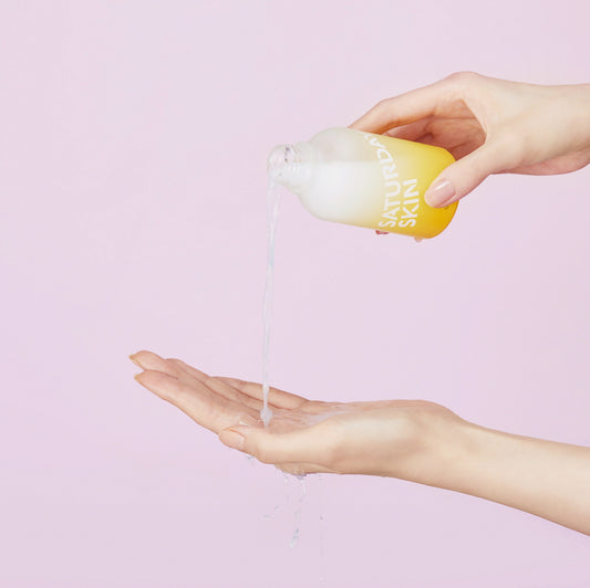 Hand pouring a clear liquid from a yellow bottle labeled 'Cure' onto another hand against a light pink background.