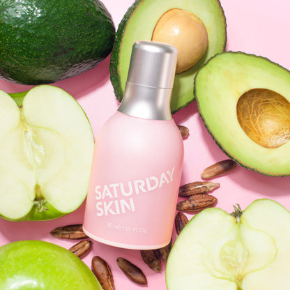Pink 'Saturday Skin' bottle surrounded by avocados and apples on a pink background